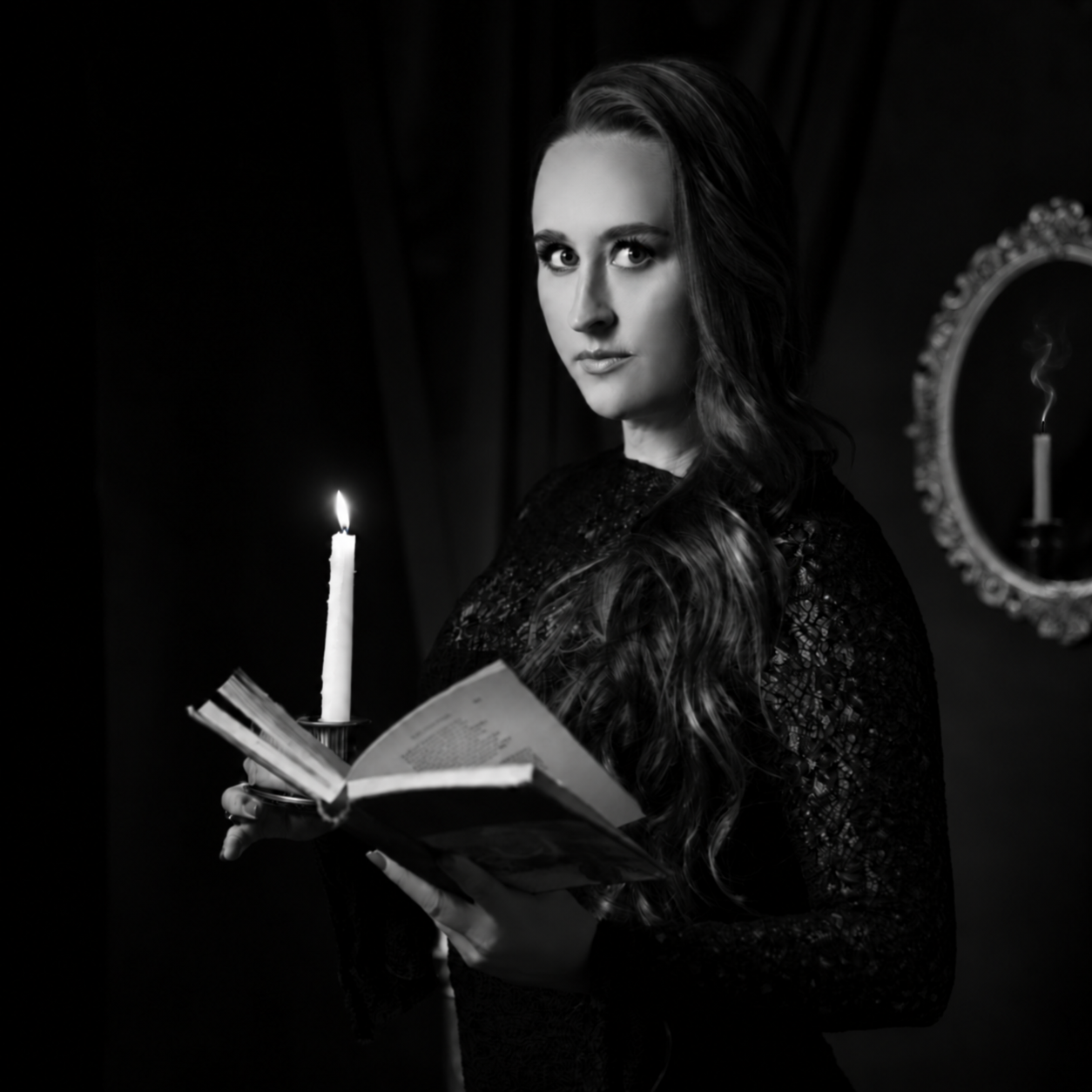 Black and white portrait of Nikki Bodie holding an open book and a lit candle in a dark room.
