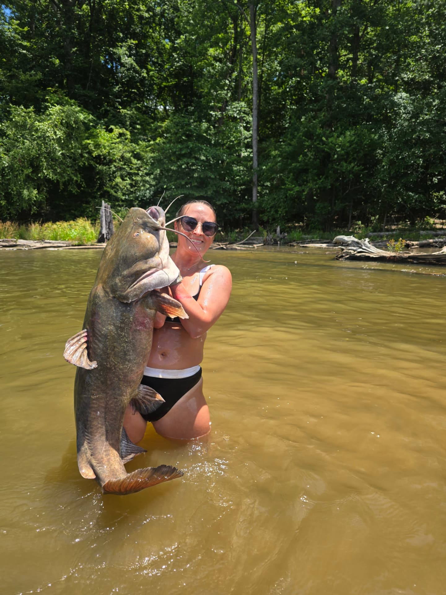 Nikki Bodie standing waist-deep in a green river holding an enormous flathead catfish.