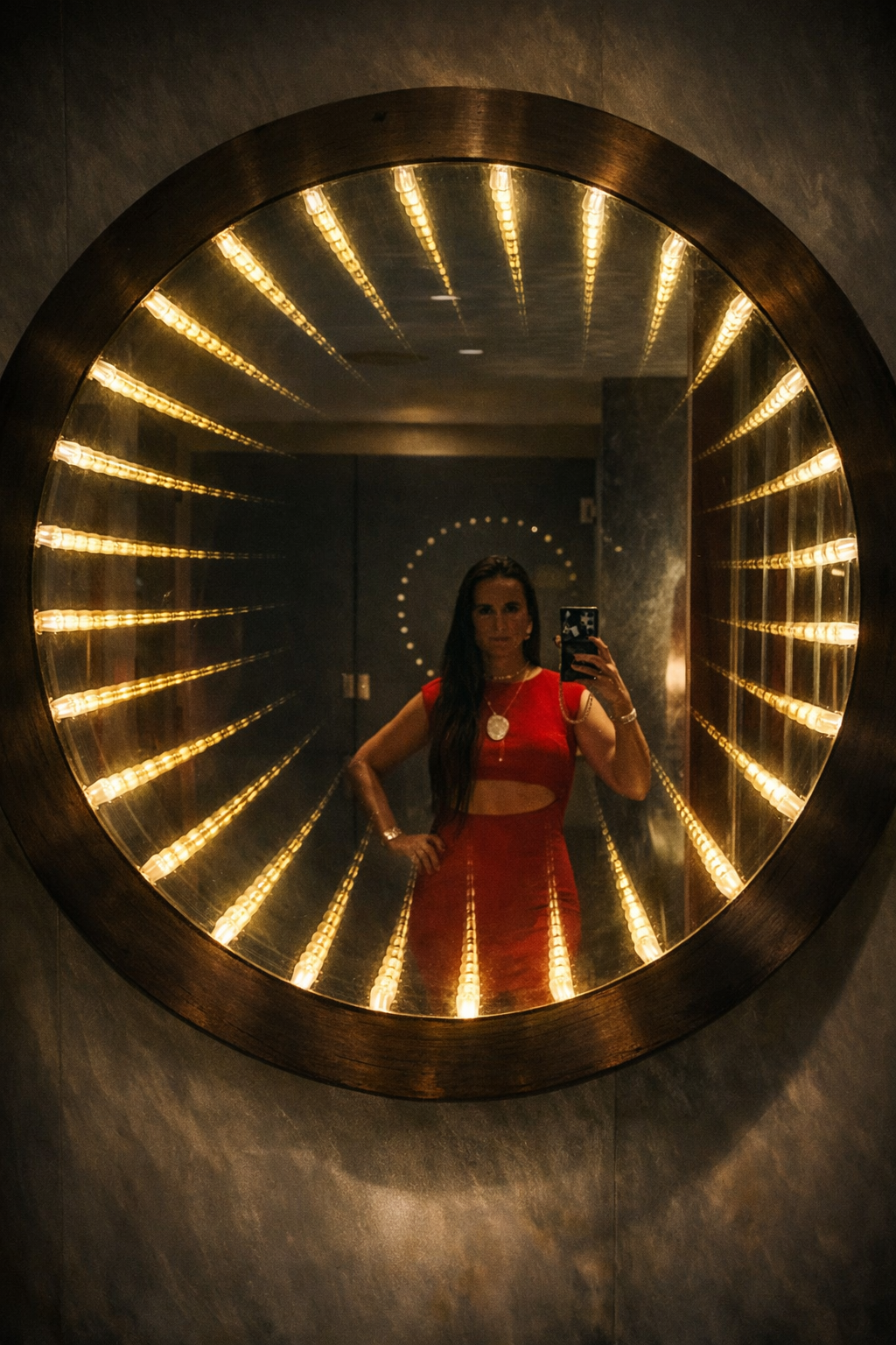 Nikki Bodie in a red dress reflected in a large round sunburst mirror ringed with warm bulbs.