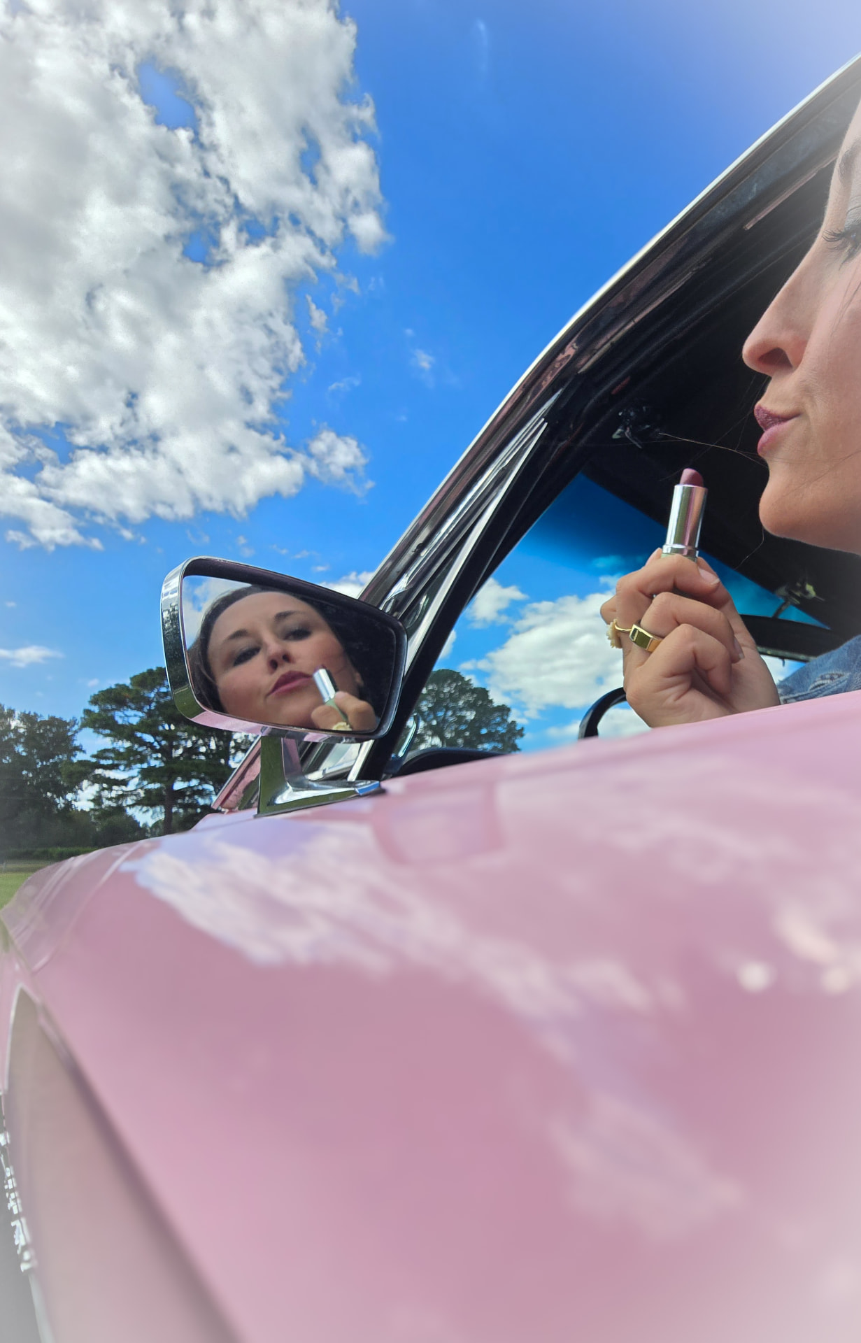 Nikki Bodie in a denim jacket applying lipstick in the side mirror of a pink Mustang under a bright blue sky.