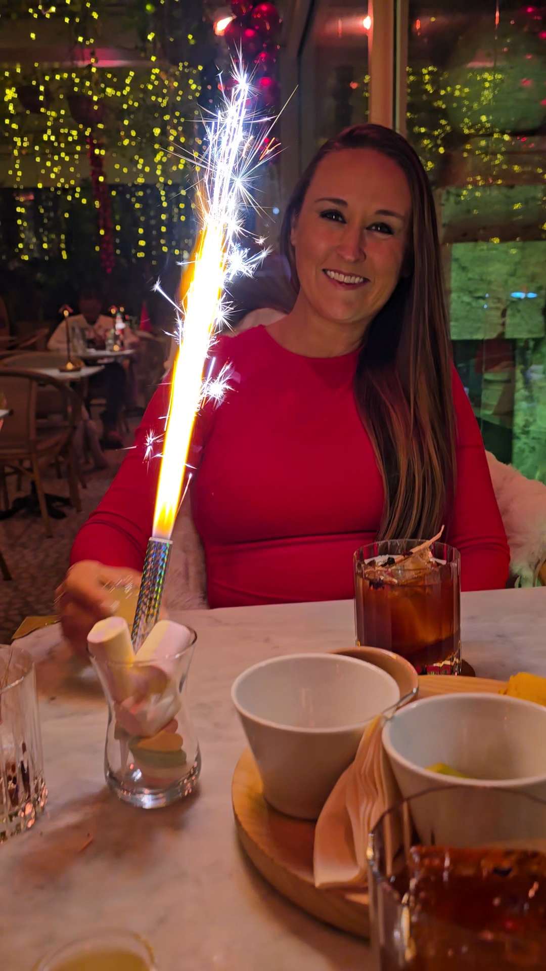 Nikki Bodie in a red dress smiling behind a lit sparkler at a candlelit table strung with fairy lights.