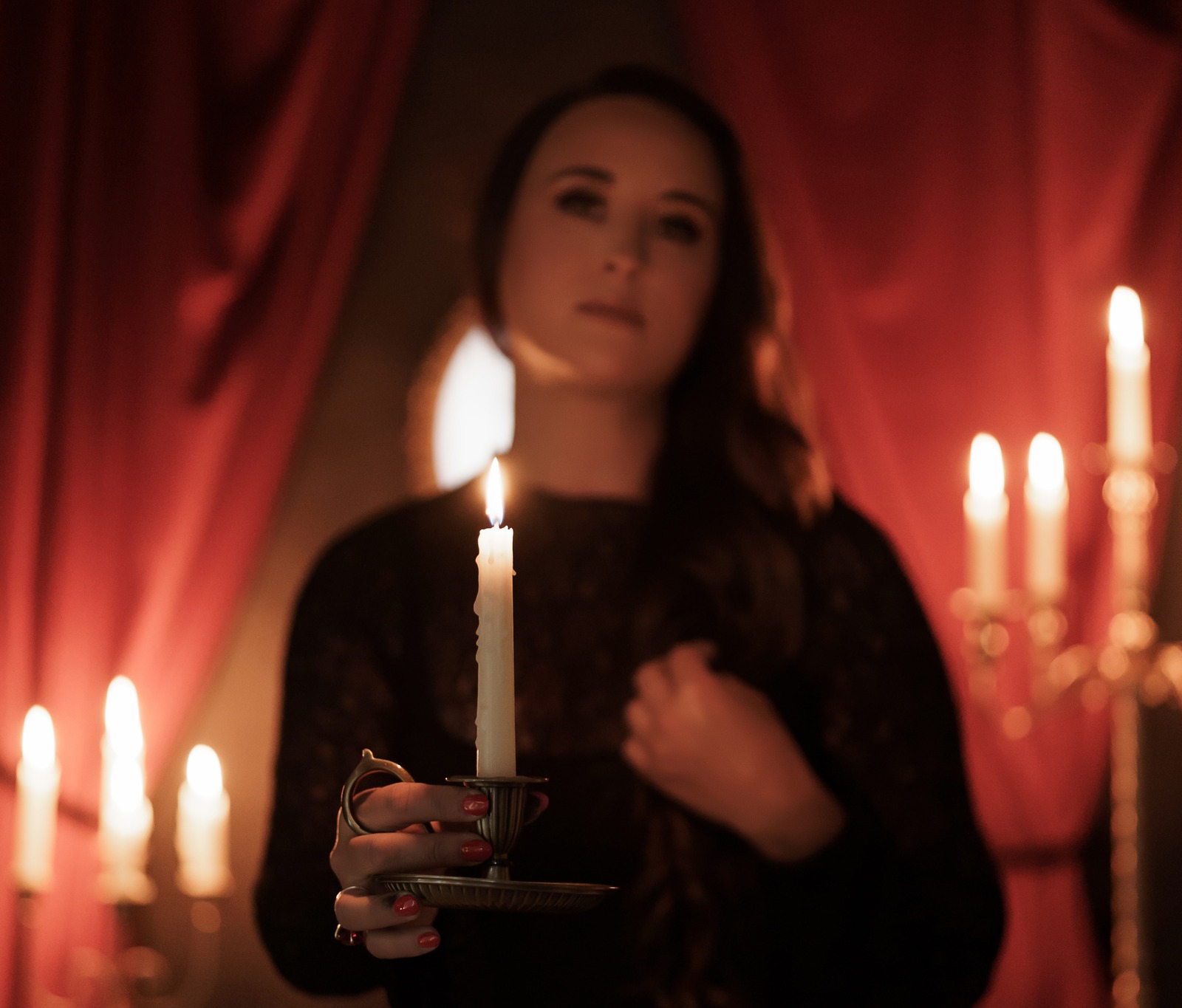 Nikki Bodie holding out a single lit candle in a brass holder before red curtains, her face soft behind the flame.