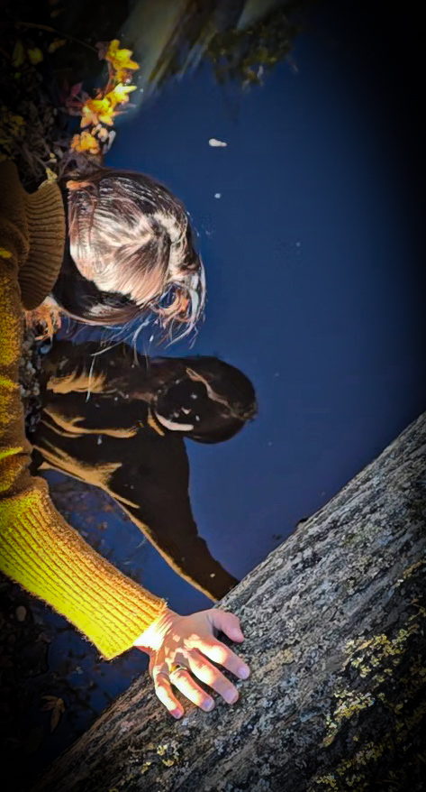 Nikki Bodie in a mustard sweater leaning over dark water with her reflection visible on the surface.