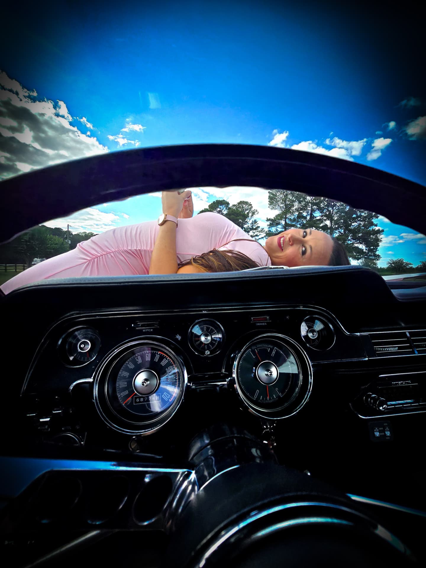 Nikki Bodie seen over the dashboard and steering wheel of a vintage Mustang against a deep blue sky.