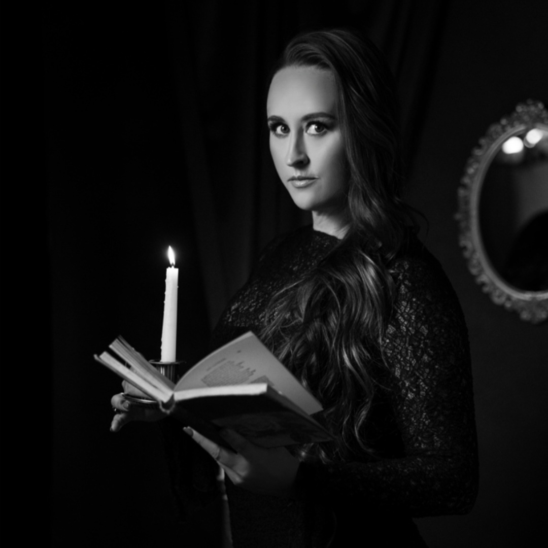 Black and white portrait of Nikki Bodie holding an open book and a lit candle in a dark room.