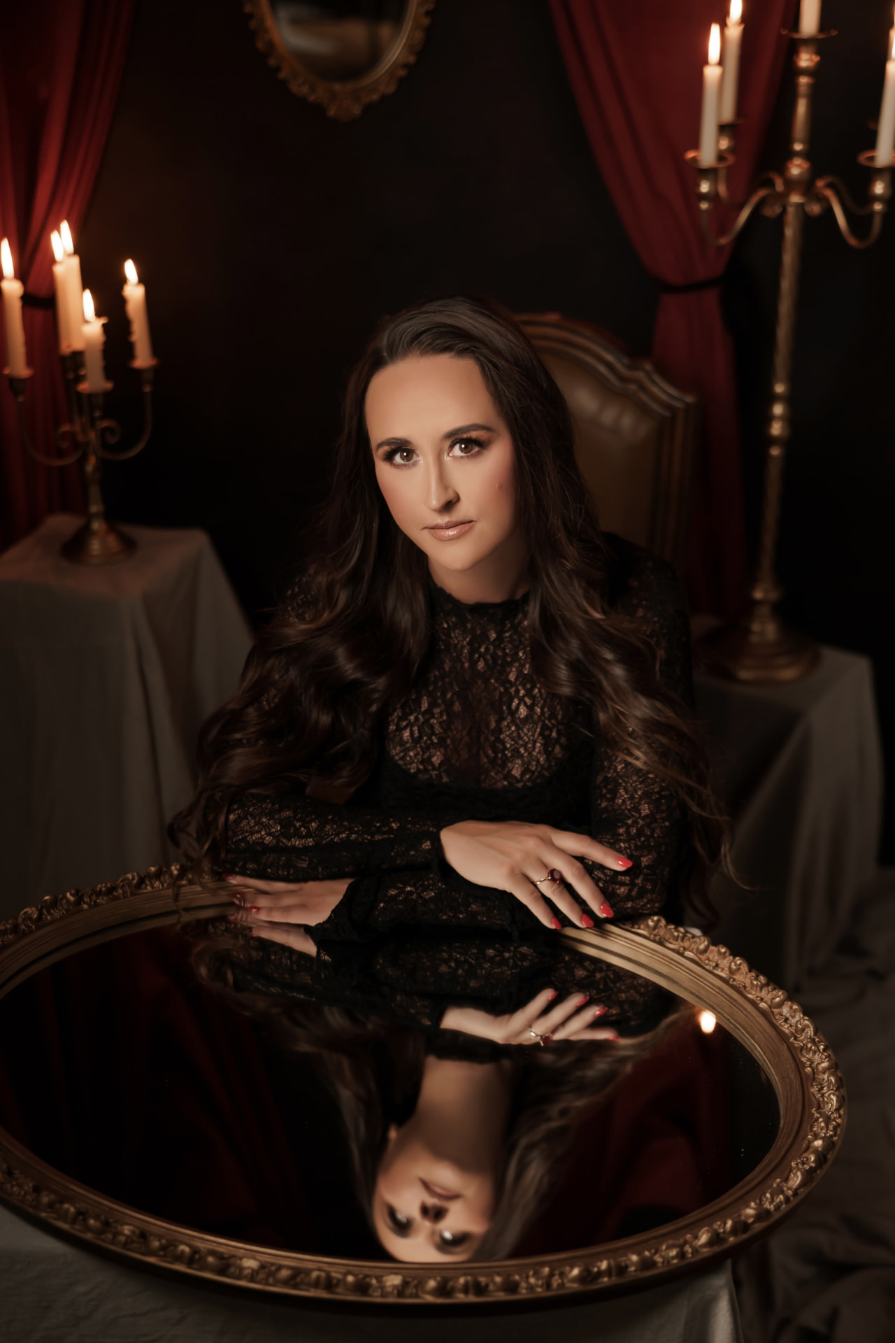 Nikki Bodie in black lace seated behind a large gilded mirror in candlelight, her face reflected upside down in the glass.